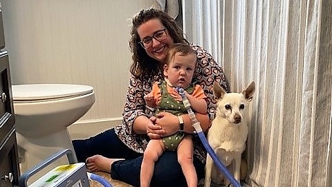 Mother smiles holding toddler with tracheostomy tube in bathroom with dog to shelter from a tornado